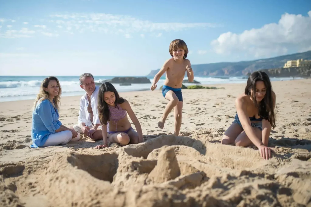 Crianças brincando na areia da praia sendo supervisionadas de perto pelos pais